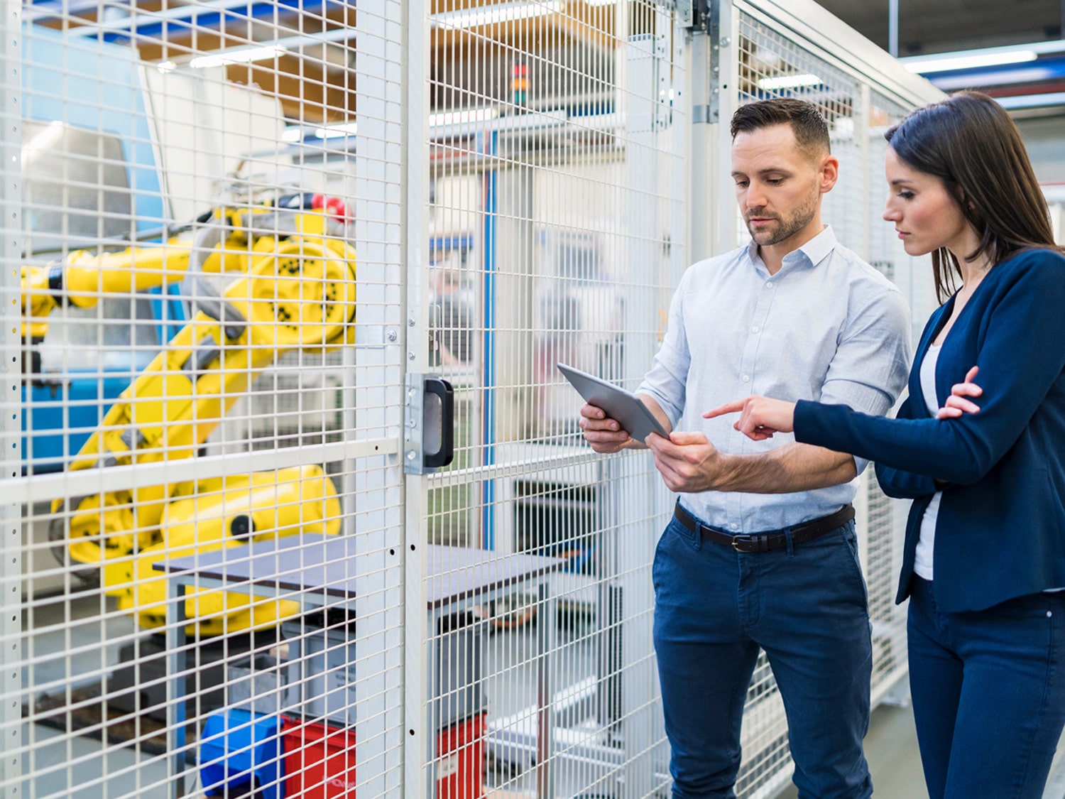 A man holding a tablet and a woman pointing to it with machinery in the background.